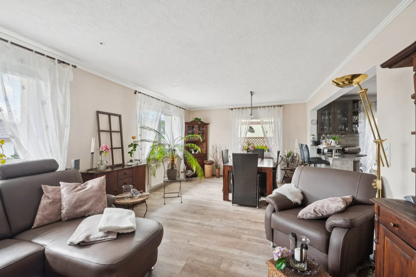 A bright living room with a brown leather sofa, armchair, dining table, and wooden furniture. Sheer white curtains adorn the windows.
