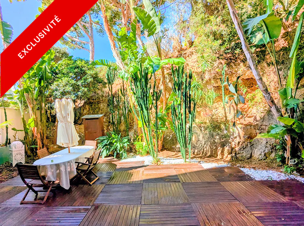 Outdoor patio with wood tile flooring, table with white cloth, chairs, and lush green plants. "Exclusivite" banner in red.