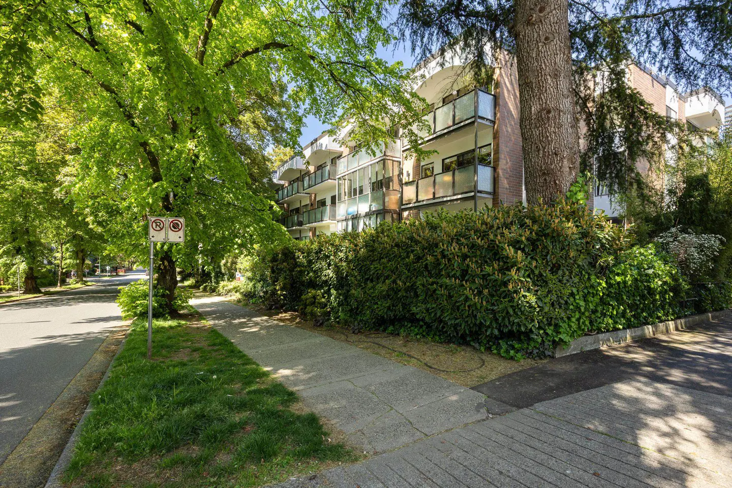 Exterior view of a multi-story apartment building with balconies, surrounded by lush green trees and bushes on a sunny day.