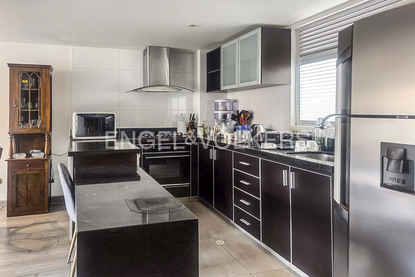 A kitchen with dark wood cabinets, stainless steel appliances, and a black countertop island. A wooden hutch stands to the left.
