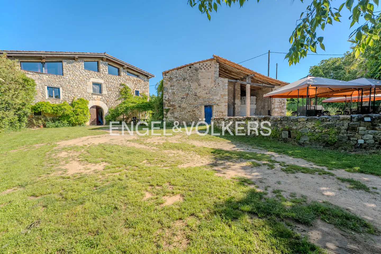 Rustic stone farmhouse with a grassy yard, an open barn structure, and orange patio umbrellas under a clear blue sky.