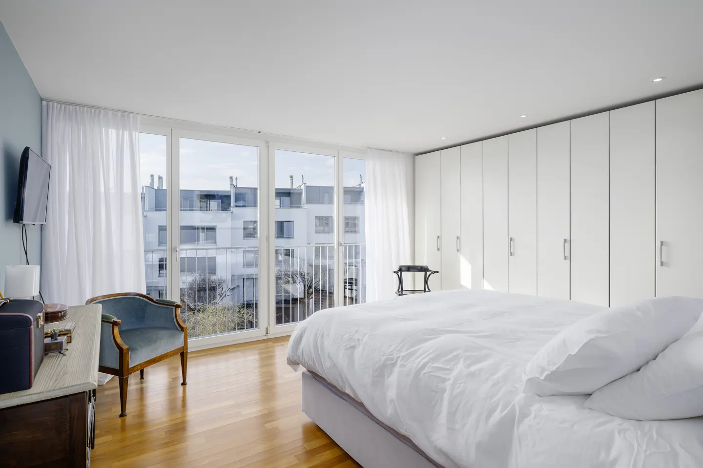 Bright bedroom with wood floors, white walls, and a large bed. Balcony doors offer a city view. A blue chair sits near a wooden dresser.