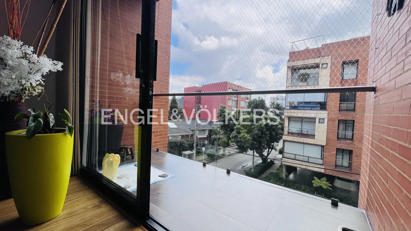 Balcony view with Engel & Völkers logo. A yellow planter sits inside. Outside, brick buildings and green trees line the street.