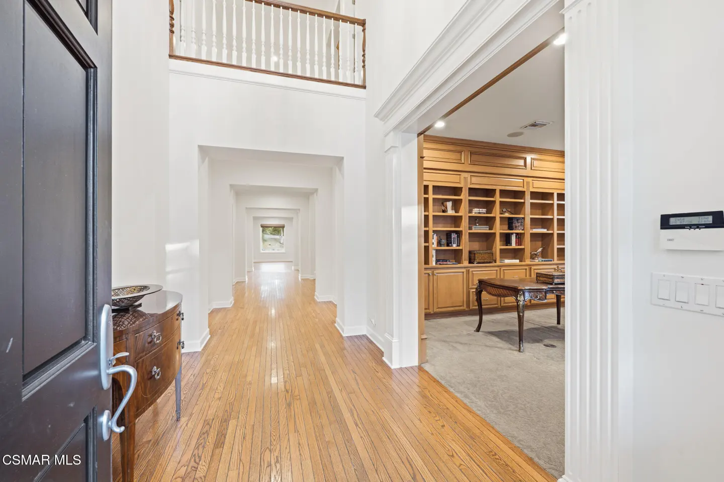 Open front door view of a bright foyer with hardwood floors, white walls, and a built-in bookcase in the adjacent room.