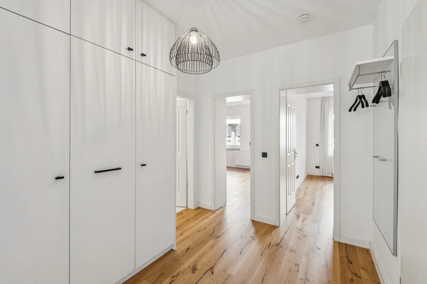 Bright hallway with white built-in closets, light wood floors, a modern wire pendant light, and a wall-mounted coat rack.
