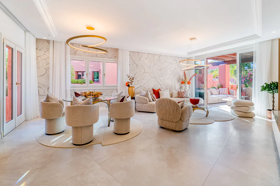 Bright, open living space with a glass table, beige chairs, and sofas. Large windows and doors lead to an outdoor patio. Modern lighting fixtures.