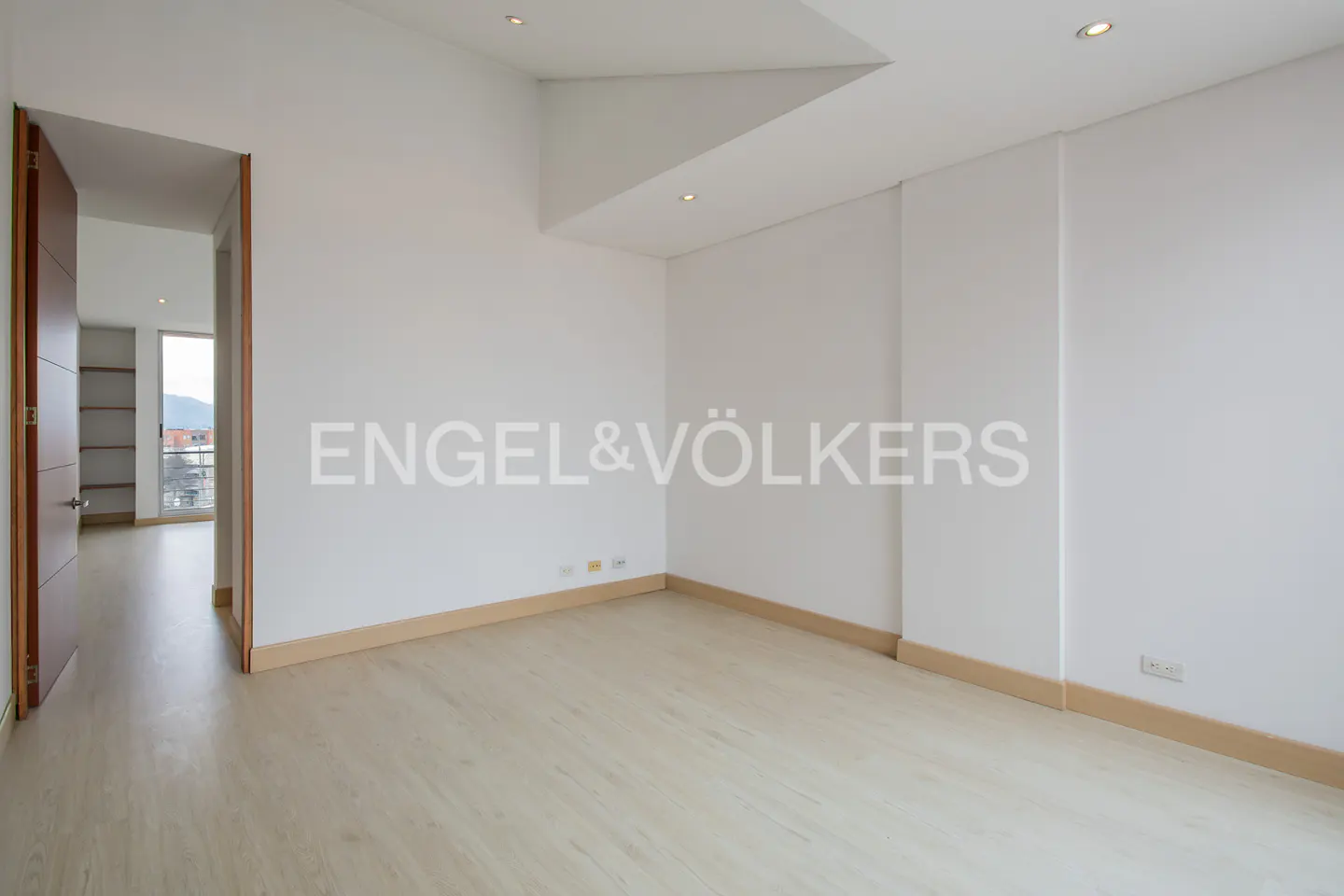 An empty, bright room with light wood floors and white walls. A doorway leads to another room with shelves and a view.