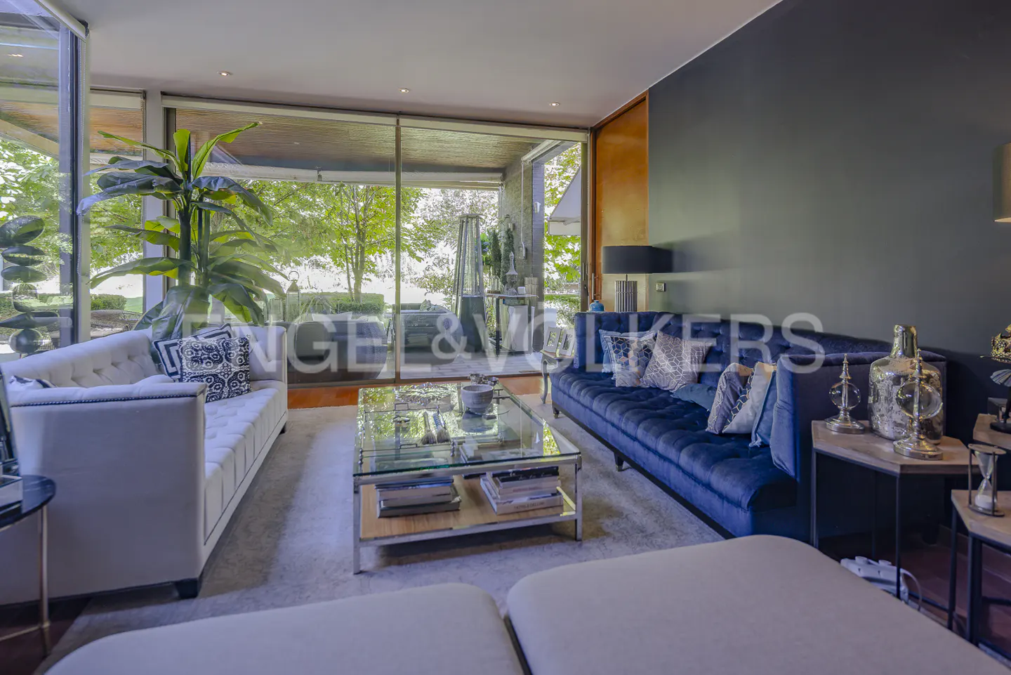 A modern living room with a blue velvet sofa, a white sofa, and a glass coffee table. Large windows overlook a green garden.