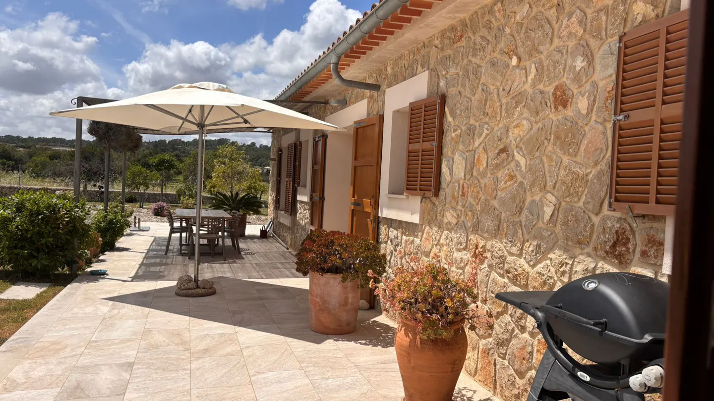Stone house exterior with a patio, featuring a dining table under an umbrella, potted plants, and a black grill.