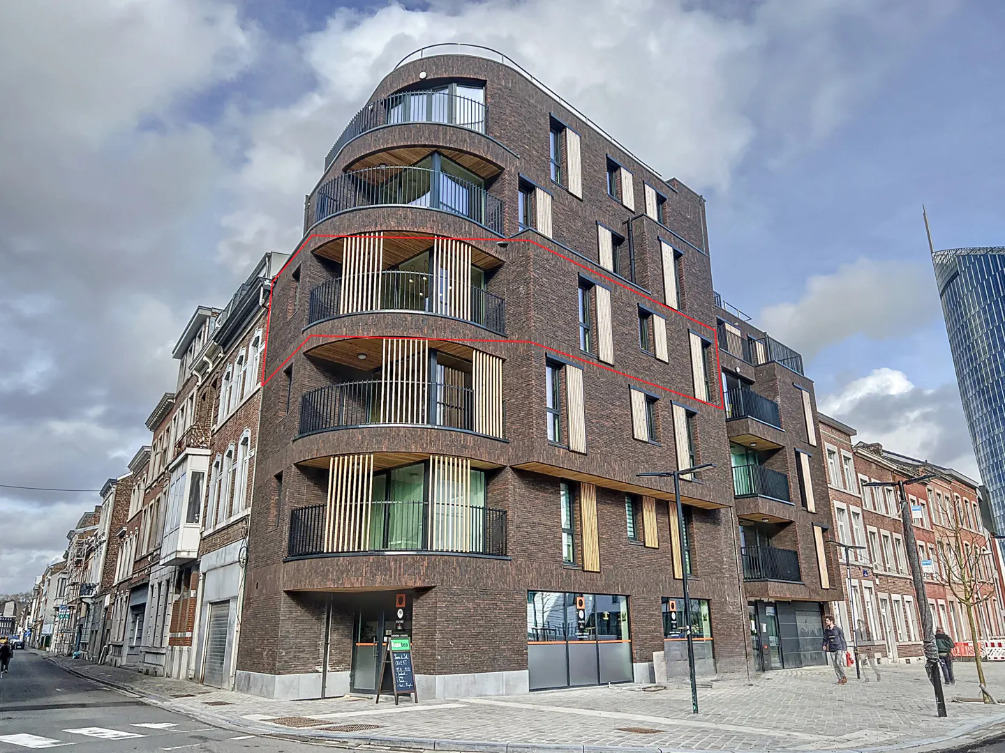 Exterior view of a modern brown brick apartment building with curved balconies and vertical wood slats. A tall glass skyscraper is in the background.