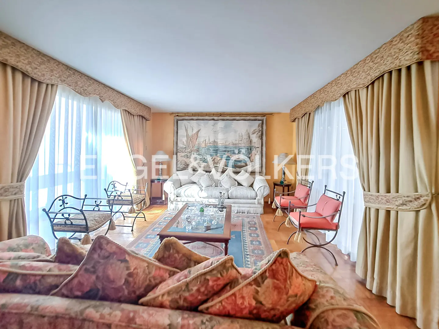A living room with floral sofas, a glass coffee table, and a tapestry on the wall. The room has tan curtains and orange walls.