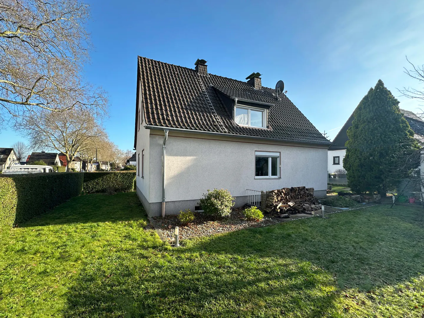 Exterior view of a white house with a brown roof, green lawn, and blue sky. A pile of firewood is stacked near the house.