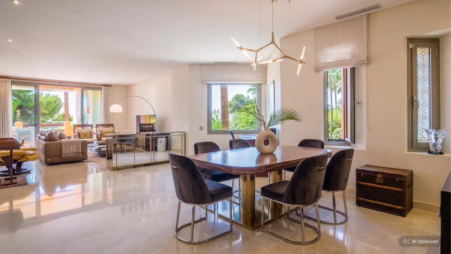 A bright, open-concept living and dining area with a wood table, velvet chairs, and a modern chandelier. A balcony and windows offer views of lush greenery.