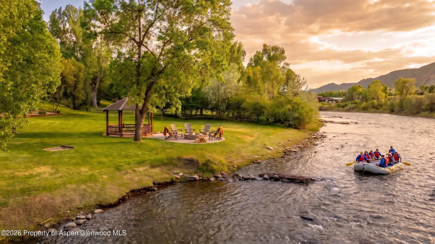 Riverfront property with raft of people paddling. Gazebo, fire pit, and chairs on the lawn. Sunset sky.