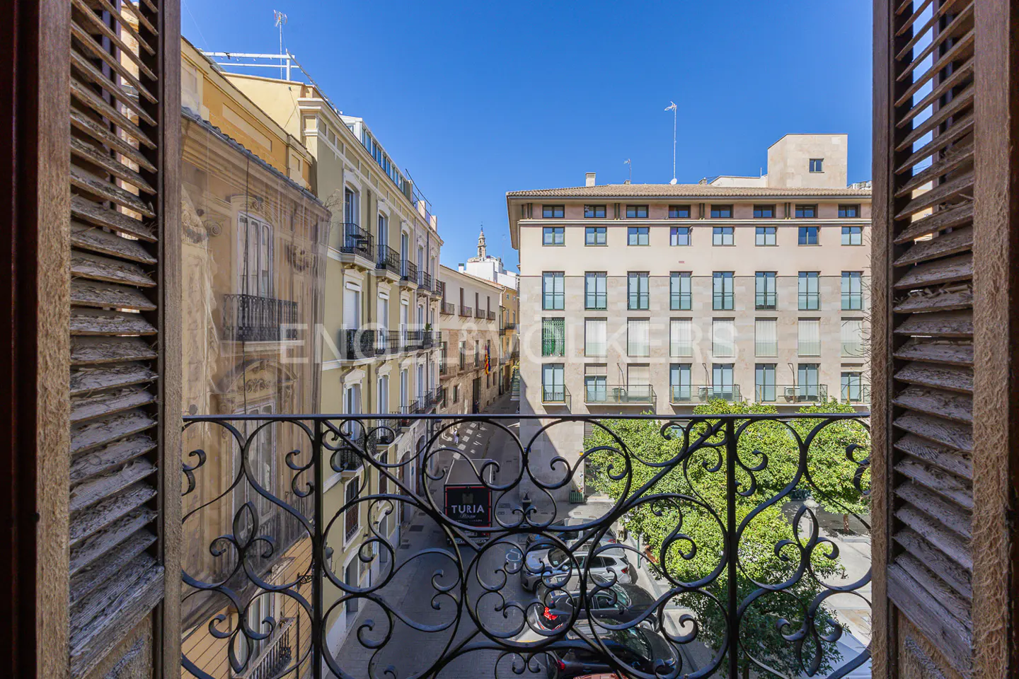 View from a window with wooden shutters and a wrought iron balcony overlooking a European city street with buildings and parked cars.