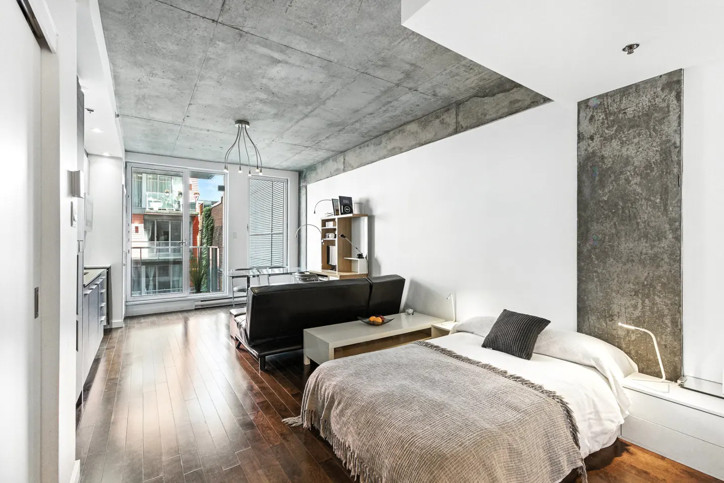 Modern studio apartment with concrete ceiling, hardwood floors, a bed, black sofa, and balcony view.