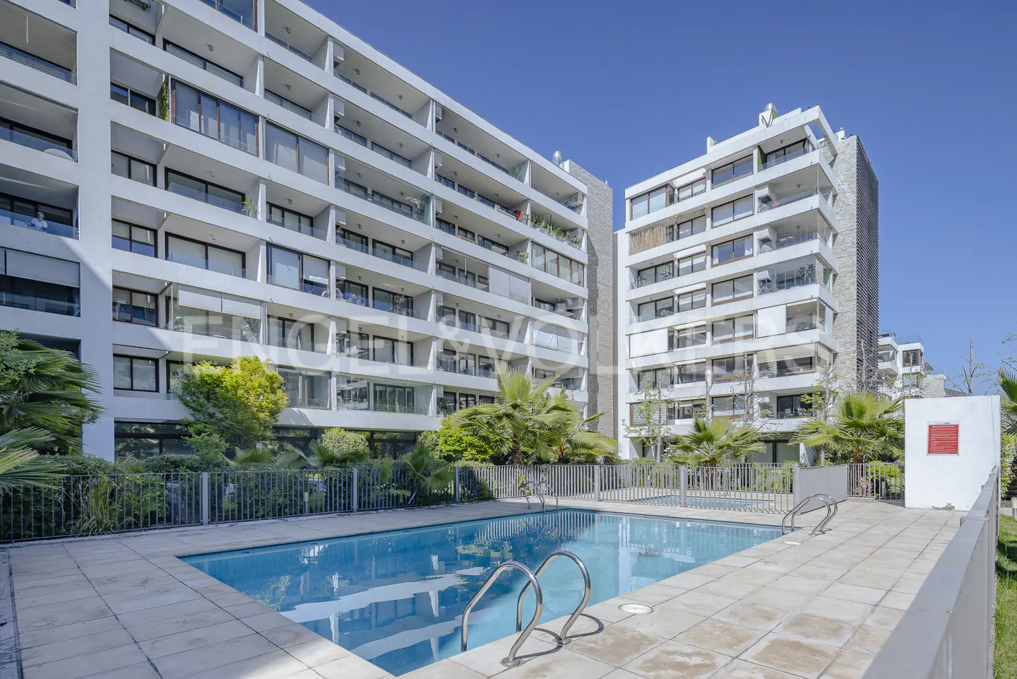 Exterior view of white apartment buildings with a blue swimming pool in the foreground.