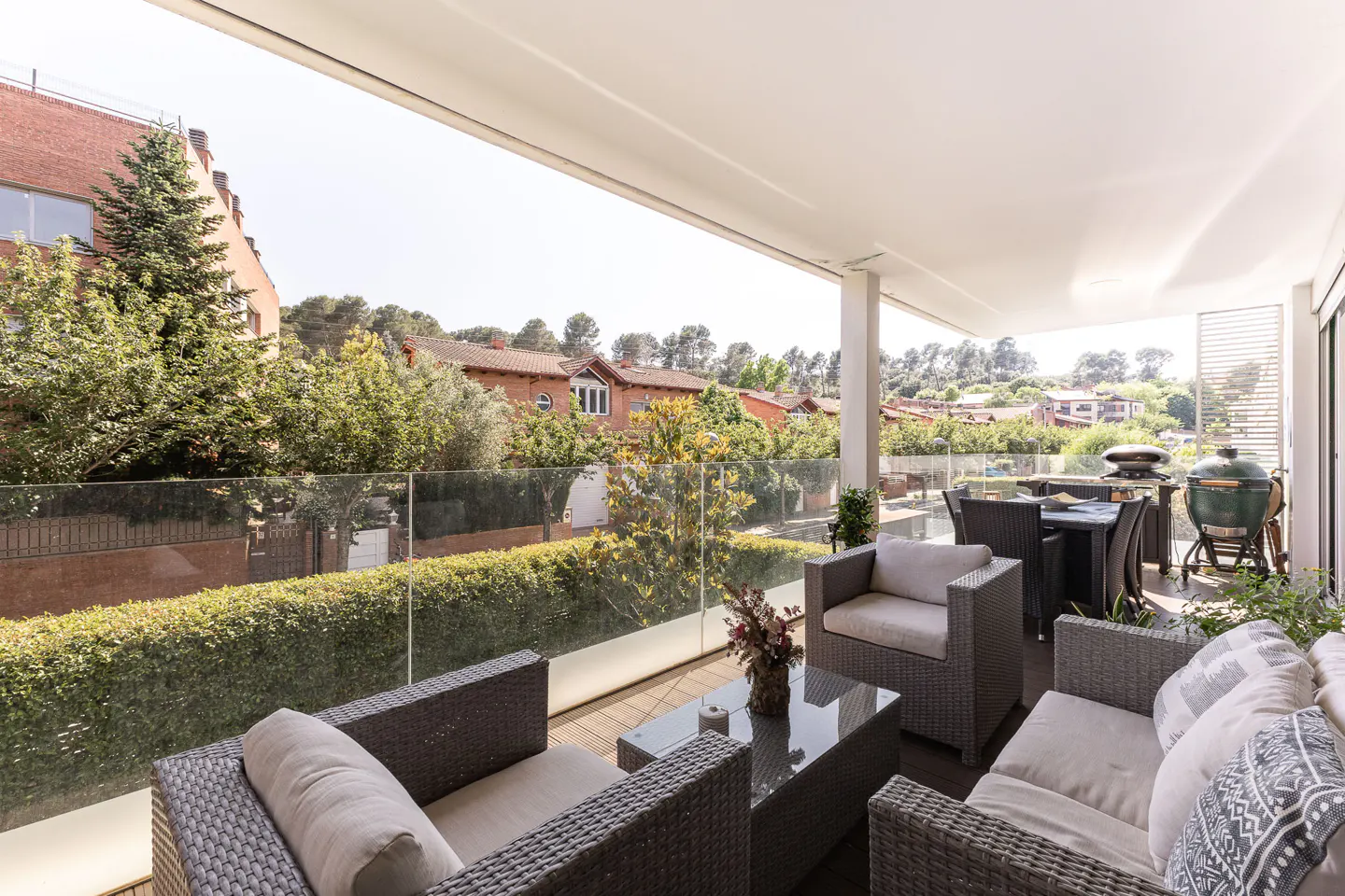 A sunlit patio with wicker furniture, including chairs, a sofa, and a glass-topped table. A grill and dining set are visible in the background, with a view of trees and houses.