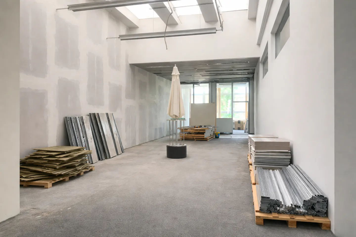 A bright, unfinished commercial space with construction materials, white walls, and a closed white umbrella in the center.