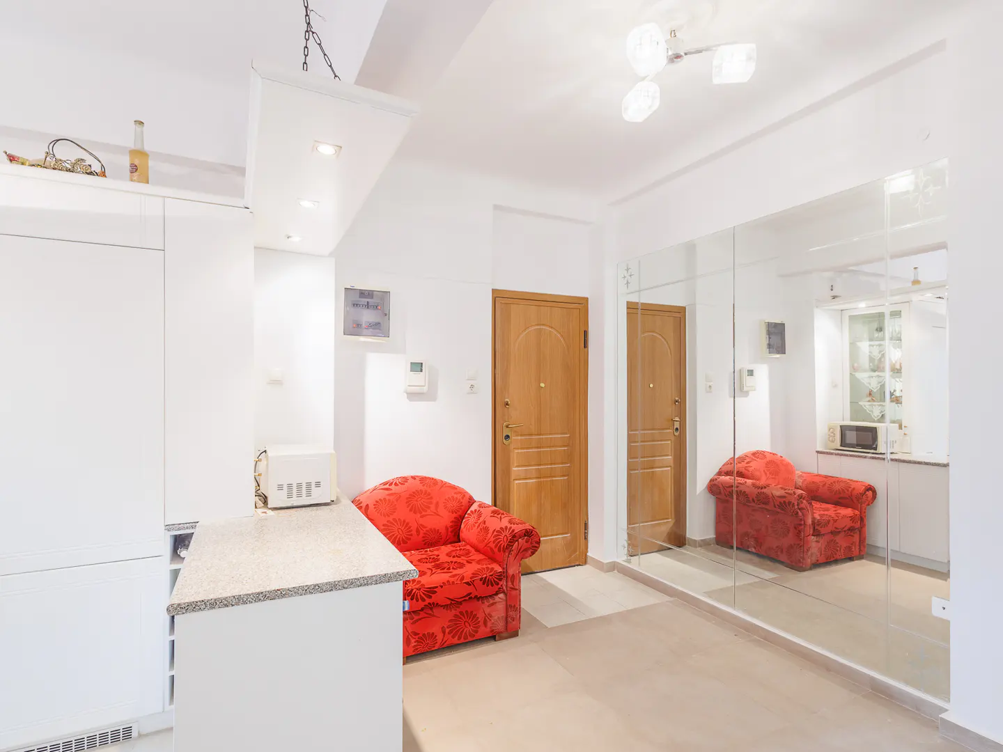 A bright room with white walls, a red patterned armchair, a wooden door, and a large mirrored wall reflecting the interior.