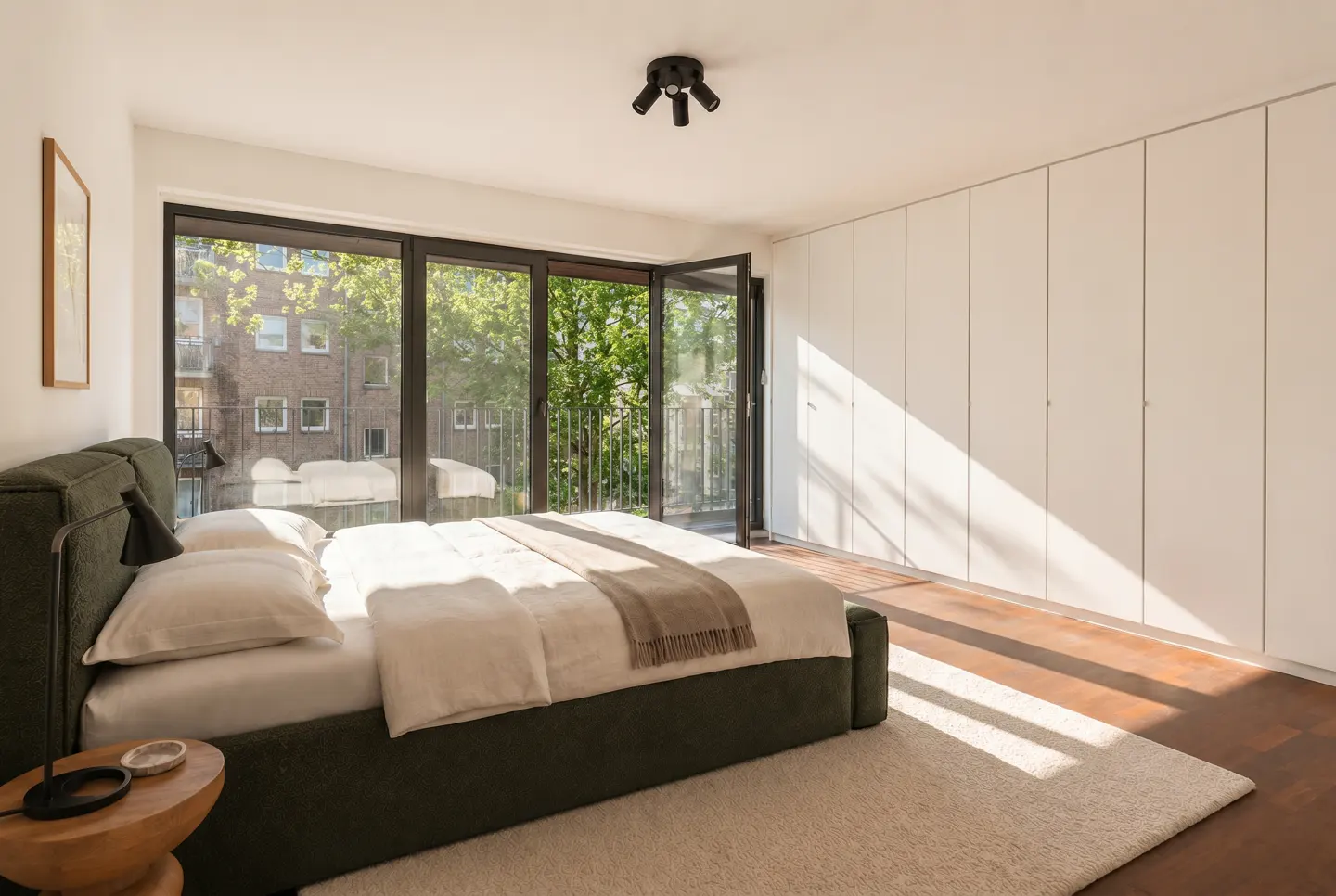 Bright bedroom with an olive green bed, white linens, and a tan throw blanket. Large windows open to a balcony with green trees.