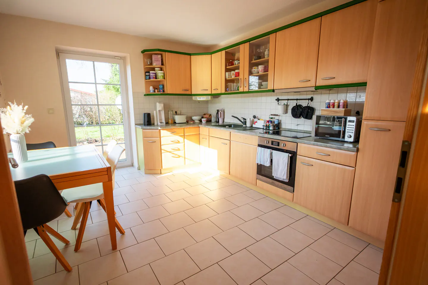 Bright kitchen with light wood cabinets, green trim, and white tile floor. A table and chairs sit near a window overlooking a garden.