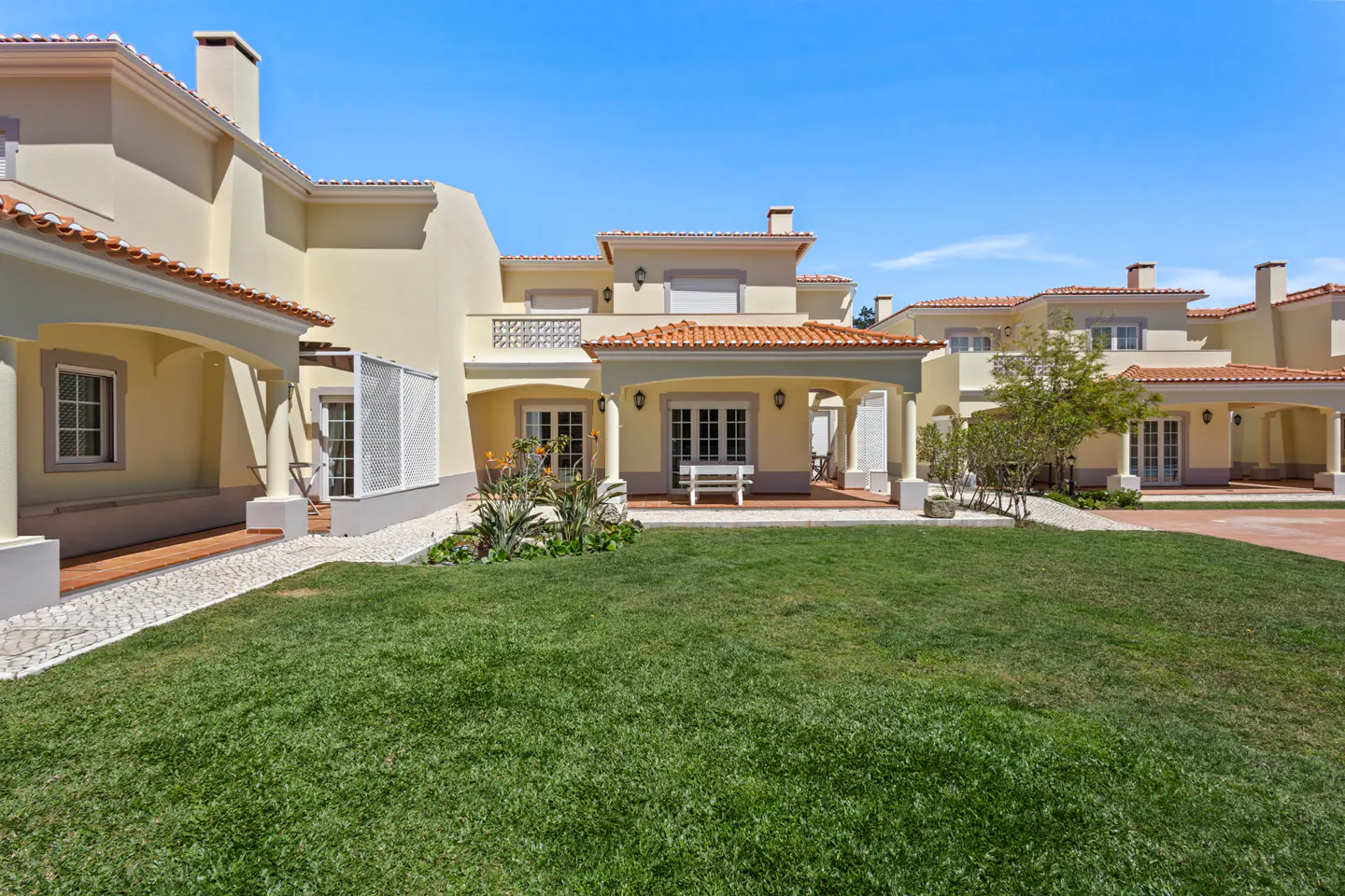 Exterior view of a light yellow house with a red tile roof and a green lawn under a clear blue sky.