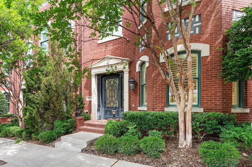 Red brick house with a decorative black door and green trimmed windows. Green bushes line the front of the house.