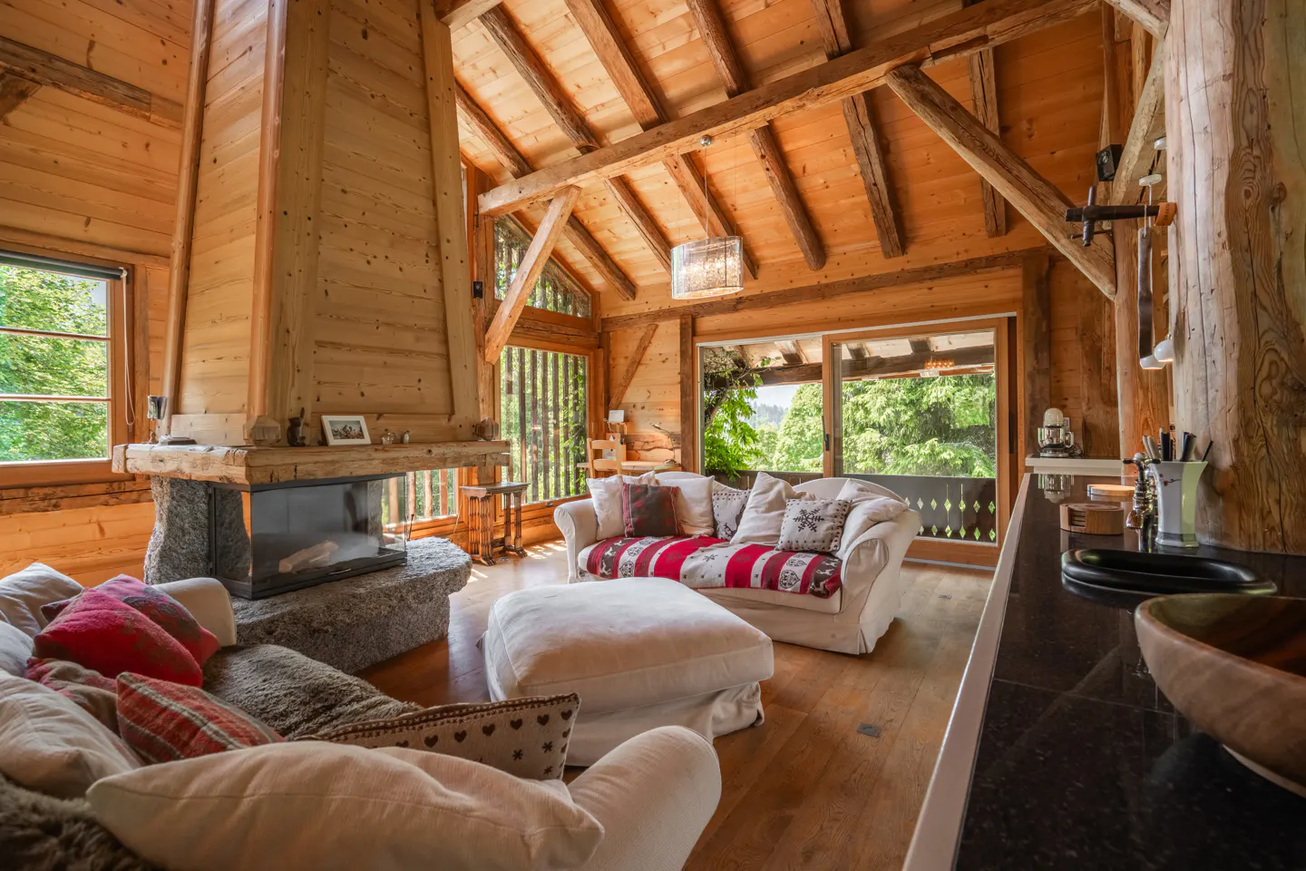 Rustic cabin interior with wood walls, exposed beams, and stone fireplace. White sofas with patterned pillows face large windows with green trees outside.