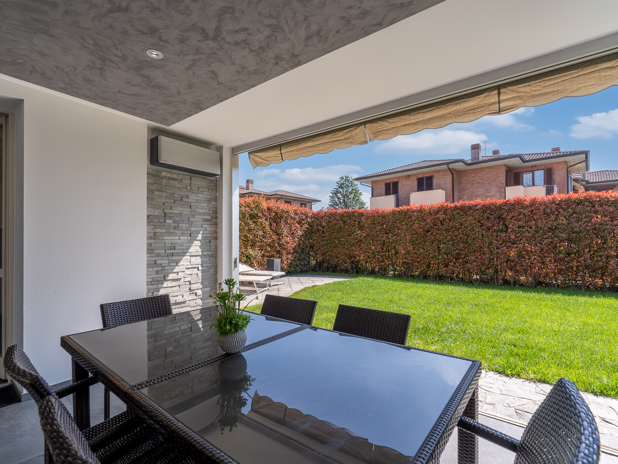 Covered patio with a glass-top dining table and wicker chairs, overlooking a private green lawn and a tall hedge. [AI-generated text]