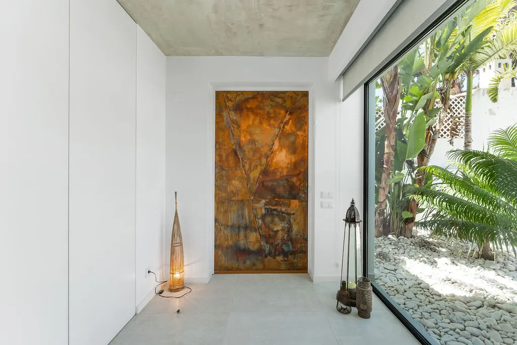 Bright entryway with a large, textured copper door, white walls, and a floor-to-ceiling window showing a lush garden.