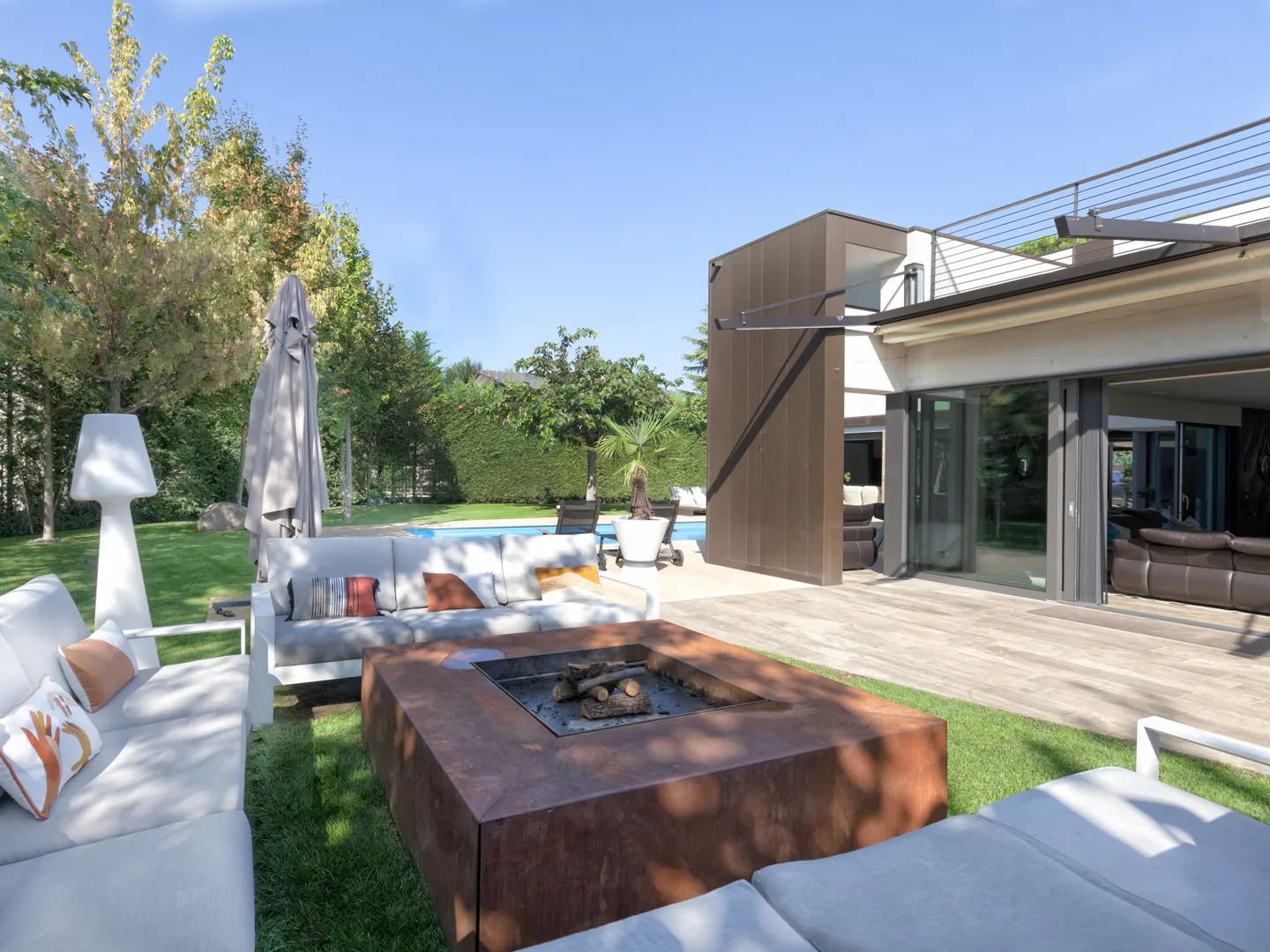 Outdoor patio with a modern fire pit, white couches, and a pool in the background. The house has large sliding glass doors.