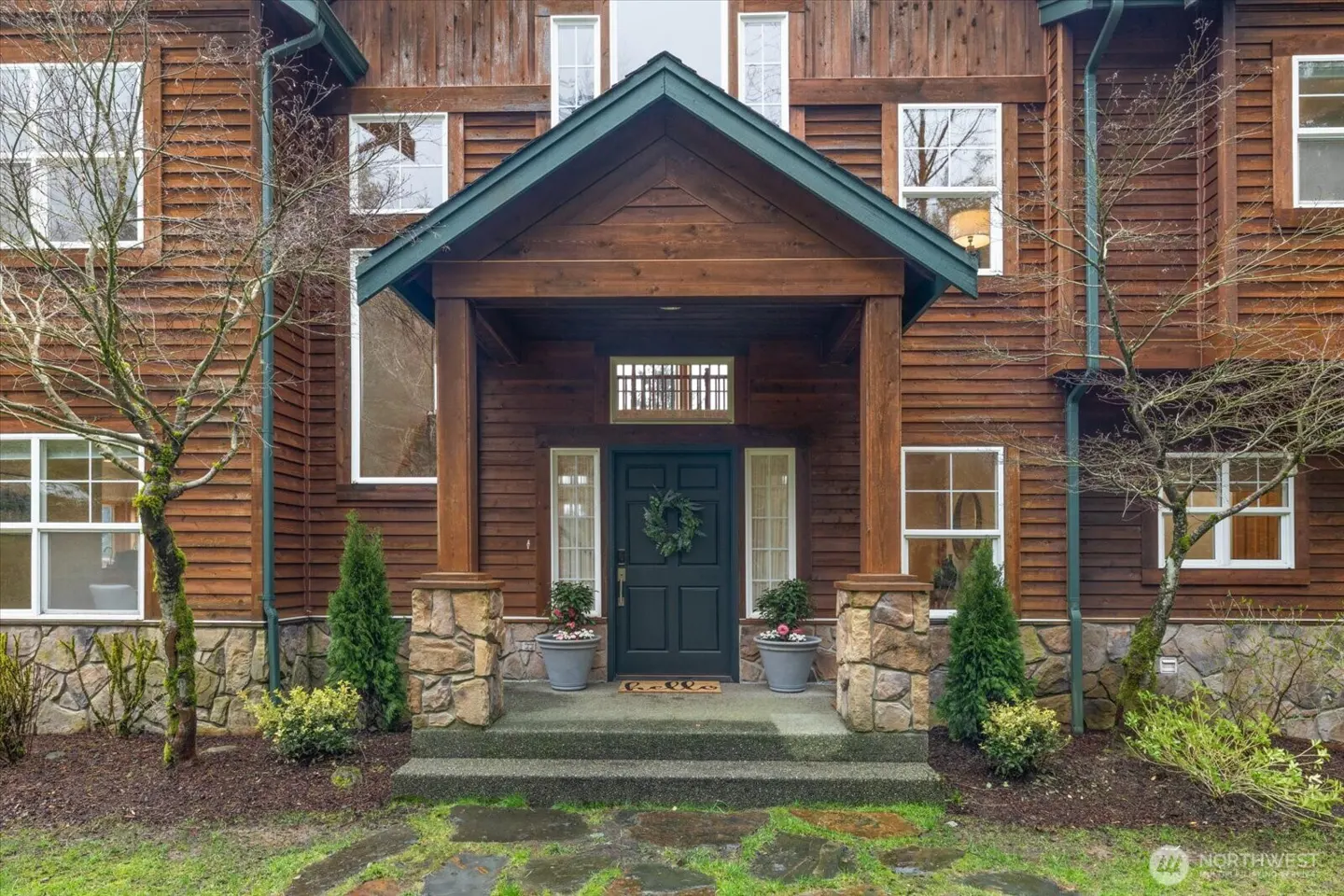 Exterior of a brown wood-sided house with a dark green front door and a wreath. Stone pillars support the porch roof.