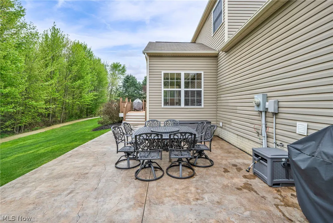 Outdoor patio with a black metal table and chairs set on a concrete patio, next to a beige house with green trees in the background.