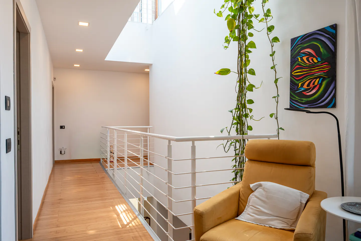 Bright hallway with wood floors, white walls, and a white railing. A yellow chair sits near a plant and colorful art.