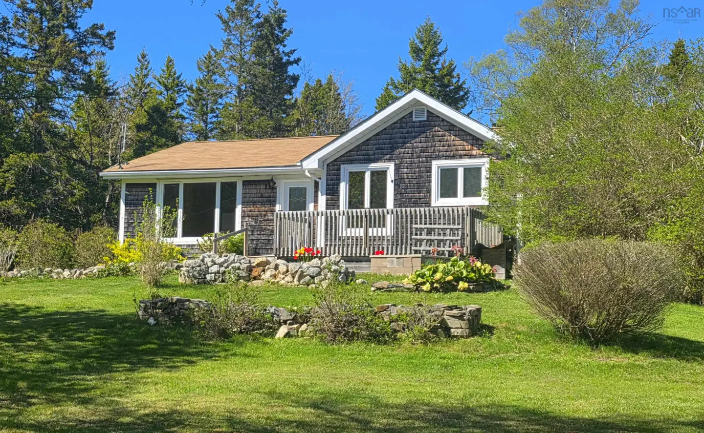 A shingled cottage with a wooden deck, surrounded by a lush green lawn, stone garden beds, and tall evergreen trees.