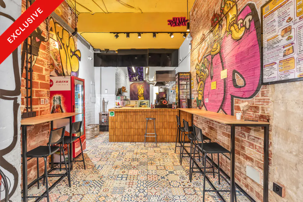 Interior view of a restaurant with graffiti brick walls, a wooden counter, and colorful patterned tile flooring.