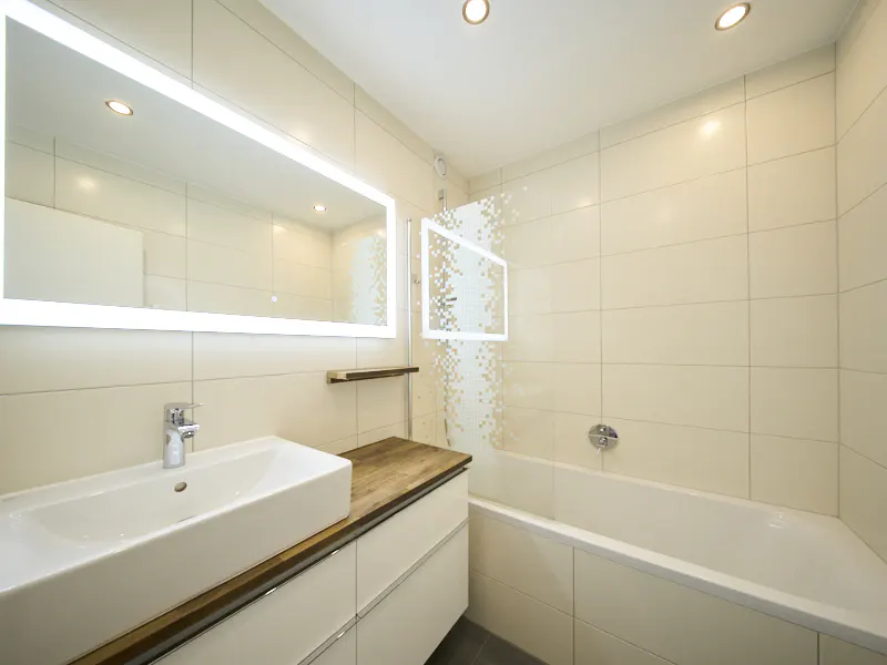 Bright bathroom with white tiles, a white tub, and a sink with a wood countertop. A large, illuminated mirror hangs above the sink.
