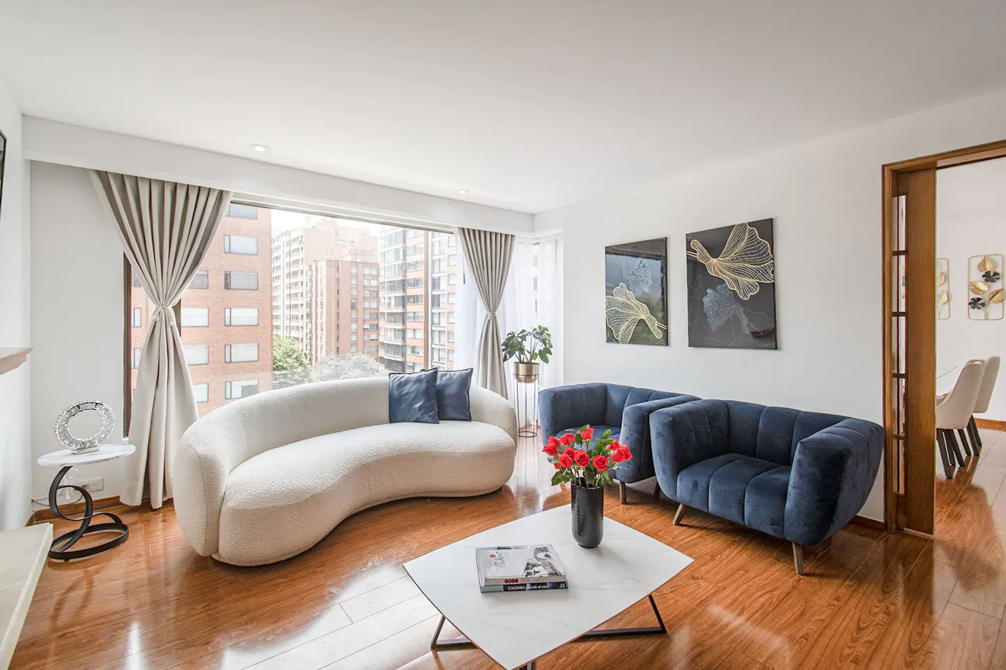 Bright living room with a curved white sofa, blue armchairs, and a marble coffee table on a wooden floor. Large window with city view.