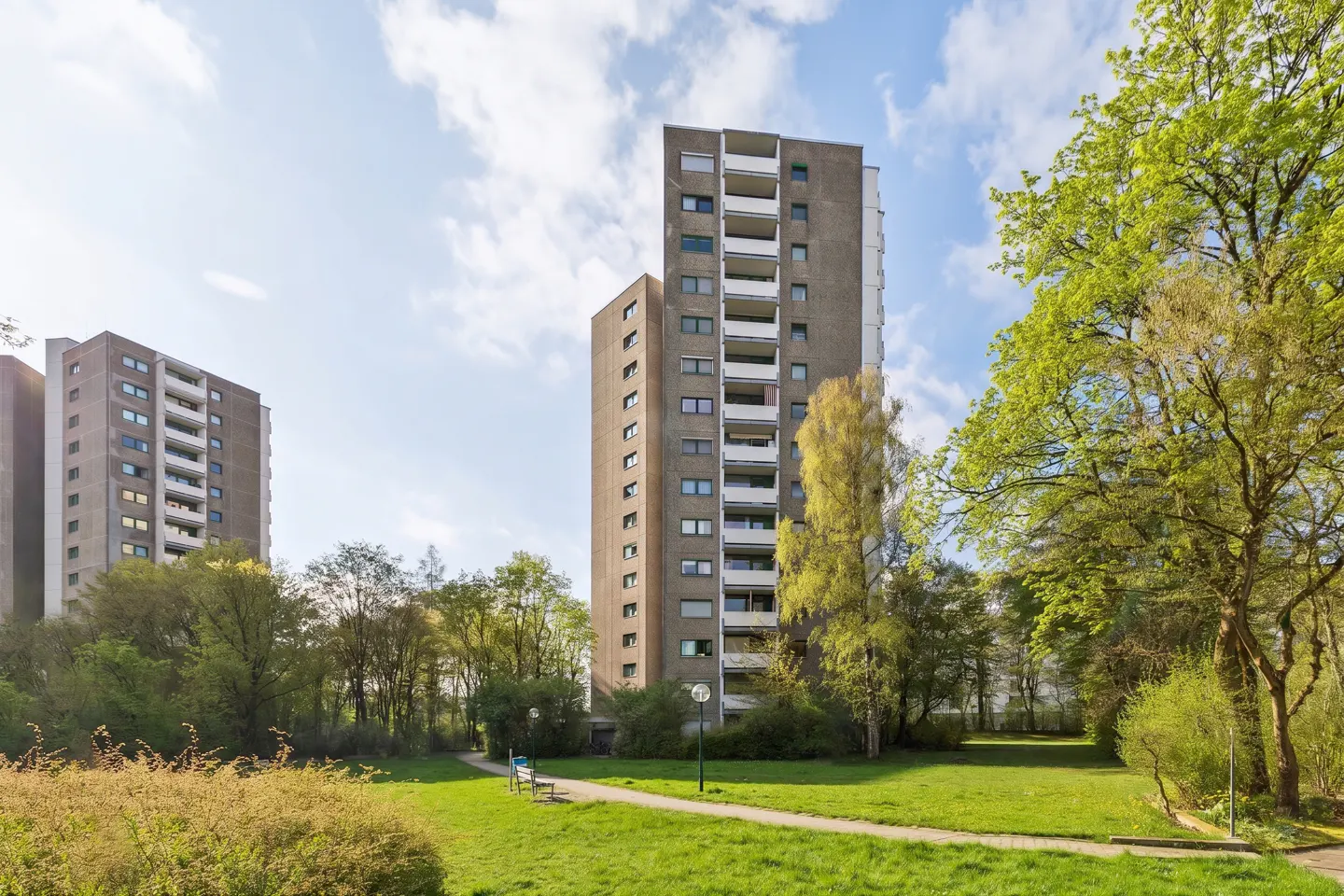 Exterior view of tall, gray apartment buildings surrounded by green trees and grass on a sunny day.