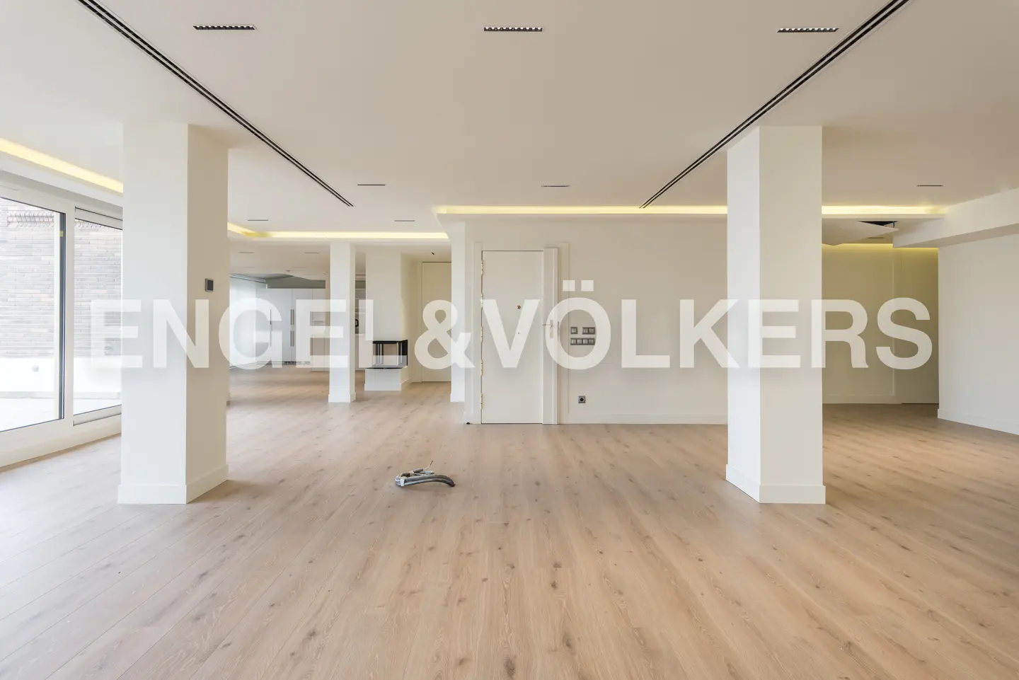 Bright, empty room with light wood floors, white walls, and support columns. A white door and fireplace are visible in the background.