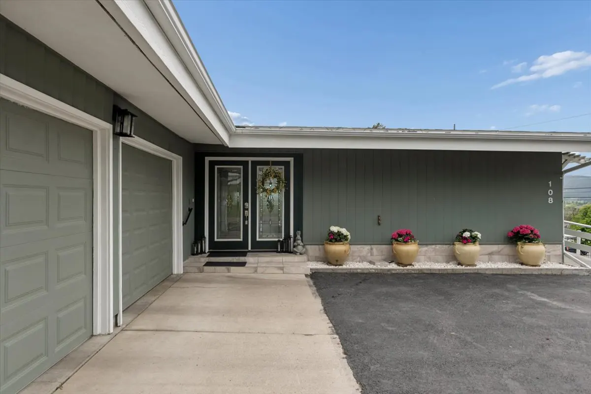 Exterior view of a one-story house with a gray garage door, olive green siding, and a black double door with a wreath. Four flower pots line the front.