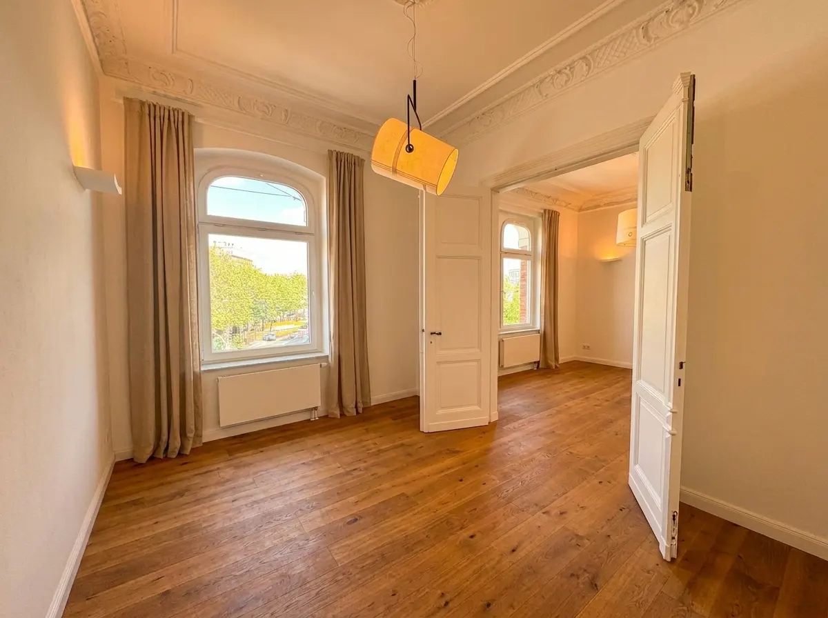 Bright, empty room with hardwood floors, white walls, and ornate ceiling trim. Two windows with beige curtains and an open white door lead to another room.