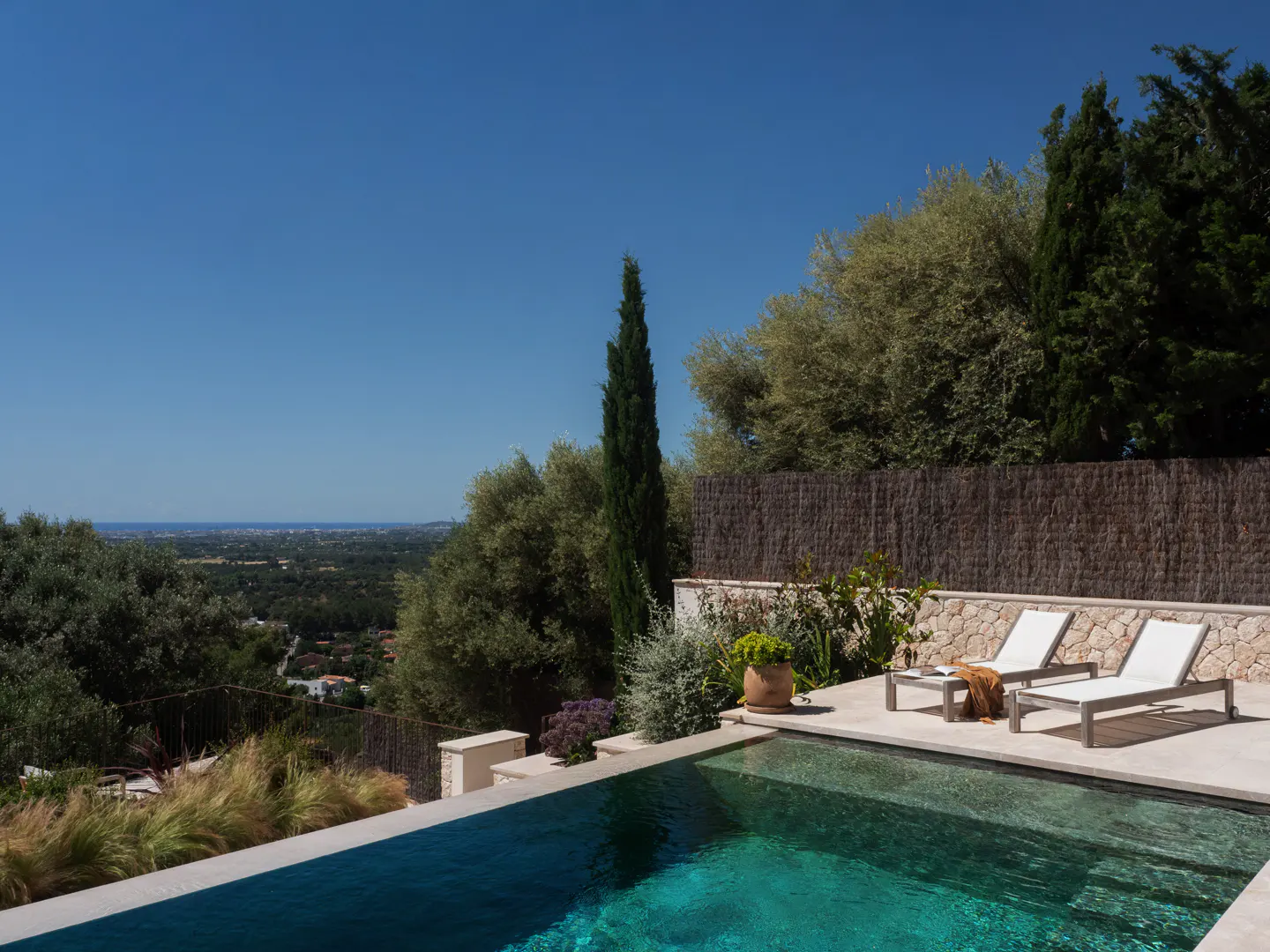 A turquoise pool with two white lounge chairs overlooks a green landscape under a clear blue sky.