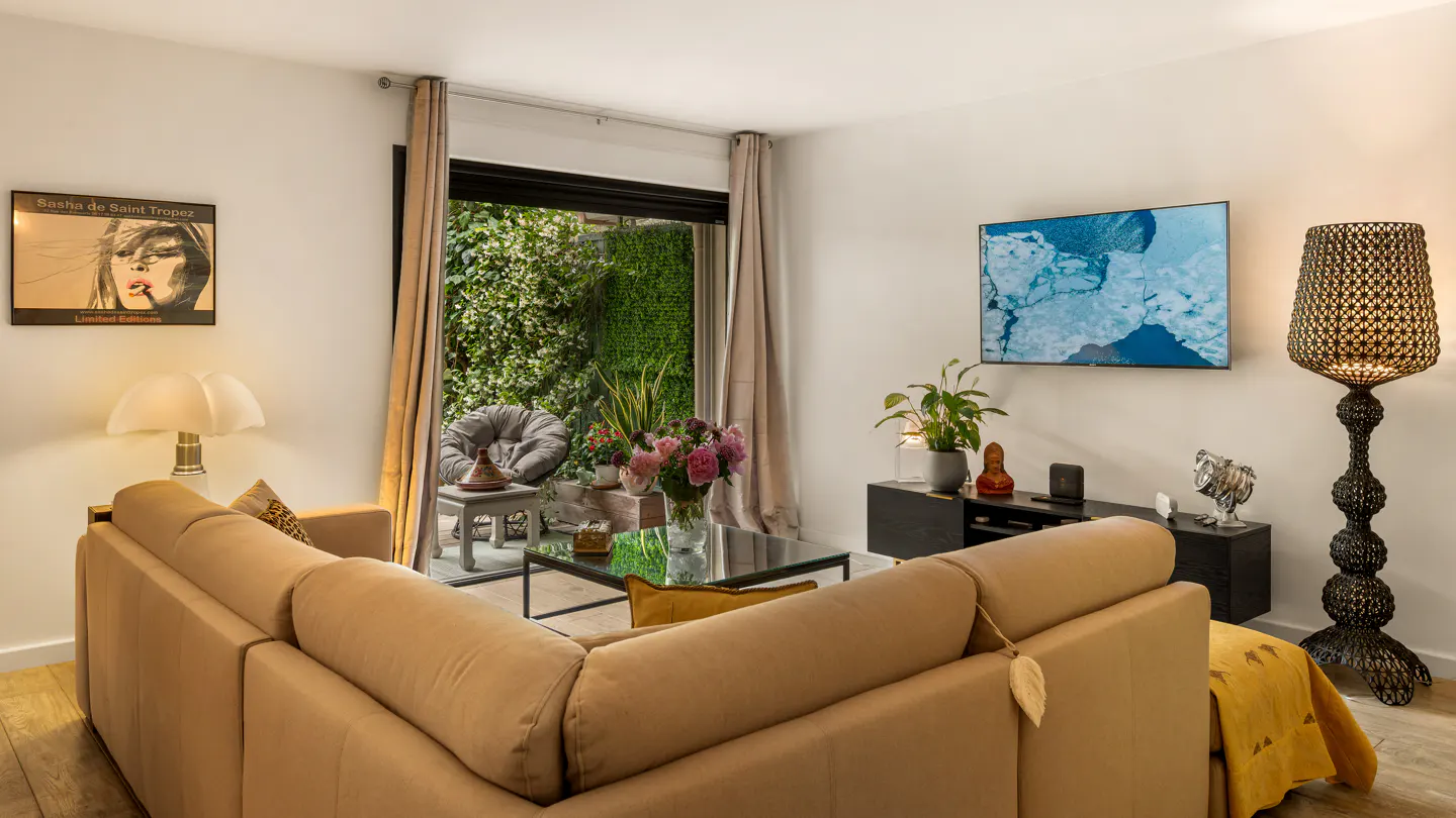Living room with a tan sectional sofa, glass coffee table, TV, and black floor lamp. A patio with green foliage is visible through a sliding glass door.