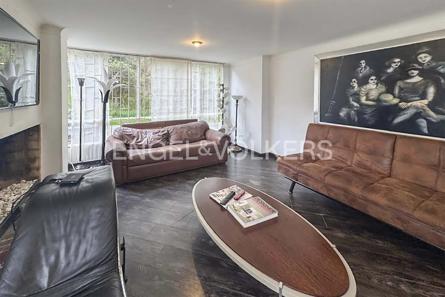 Living room with brown leather sofas, dark wood floors, and a large painting on the wall. A coffee table holds magazines. Curtains cover a window.