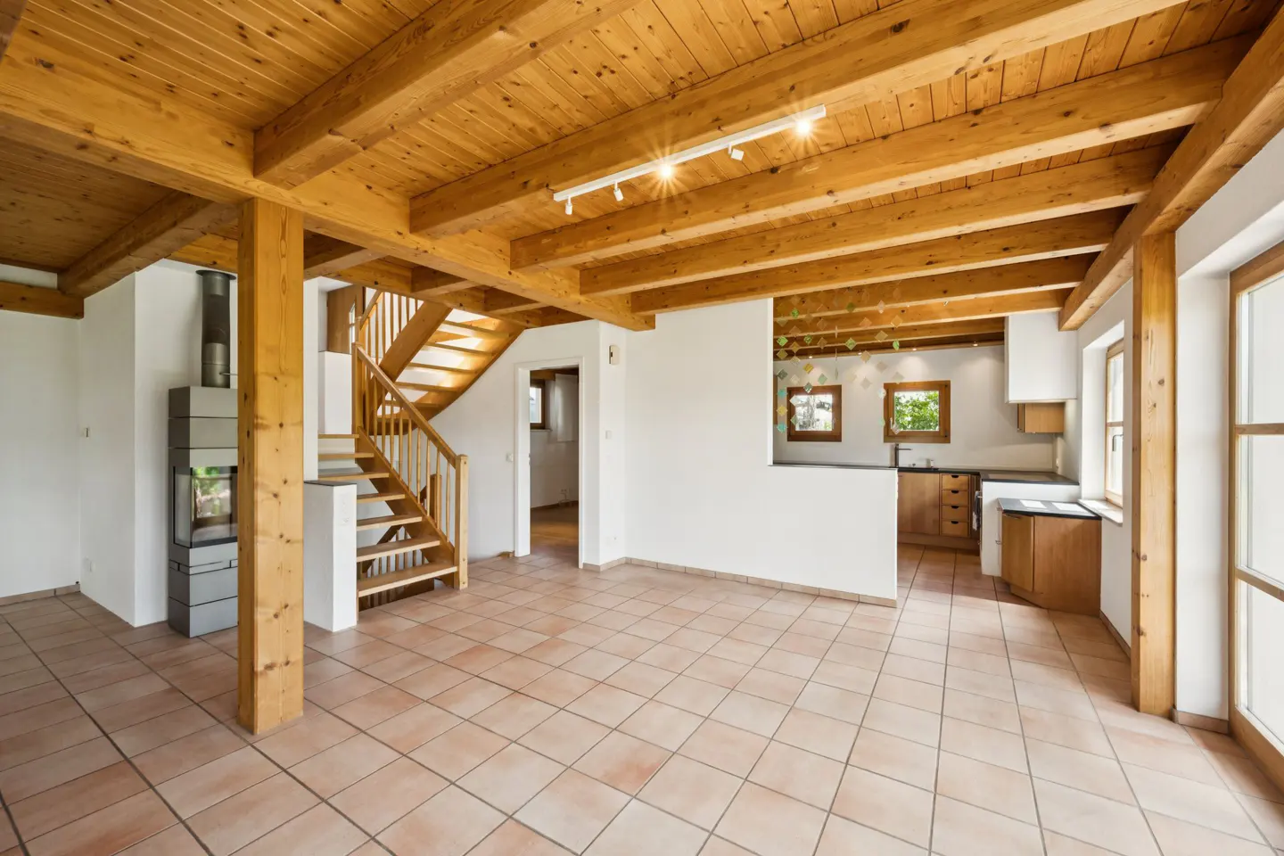 Bright, open-concept living space with wood beams, tile floor, and a modern fireplace. A staircase leads to the upper level. Kitchen in background.