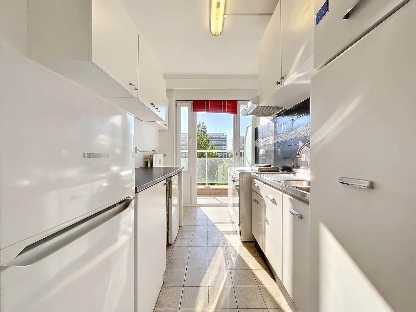 A bright, narrow kitchen with white cabinets, appliances, and tiled floor leads to a balcony with a red awning and city view.