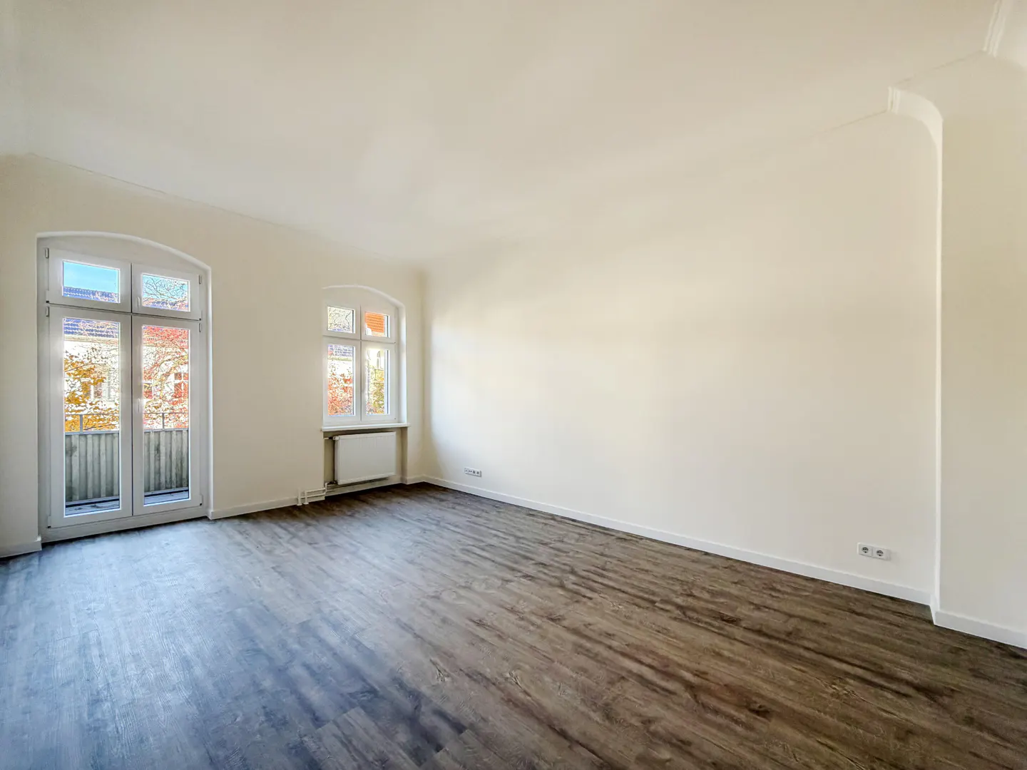 Bright, empty room with dark wood floors, white walls, and two arched windows. One window is a door to a balcony.