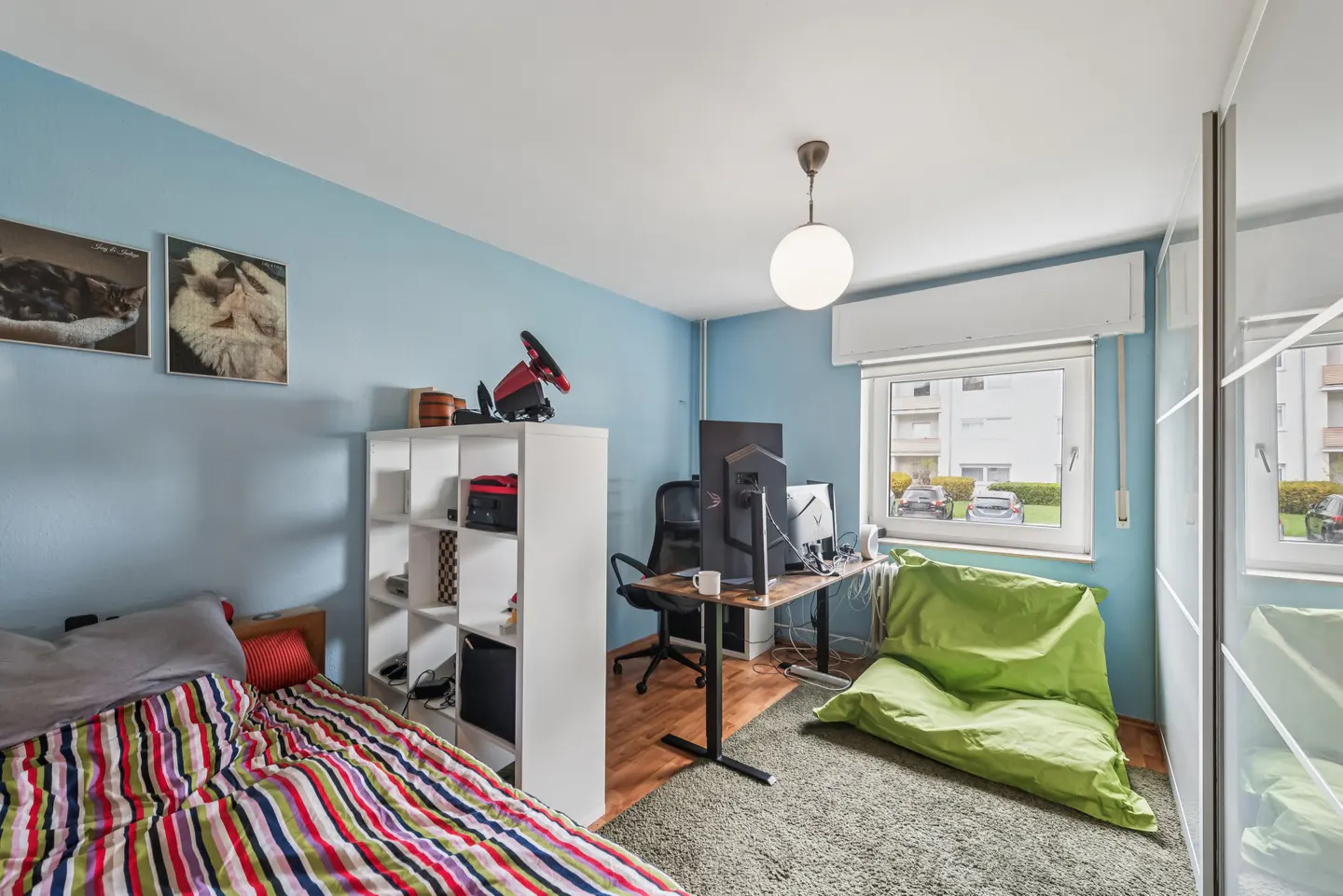 Bedroom with blue walls, a striped bedspread, a white bookshelf, a desk with computers, and a green beanbag chair.